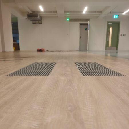 Low-angle view of newly installed wood-effect flooring with two recessed metal floor grilles in an open workspace.