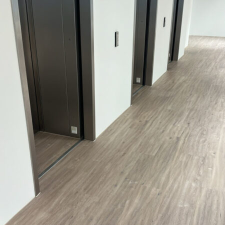 Wood-effect flooring laid along a corridor outside lift doors with white walls and dark lift surrounds.