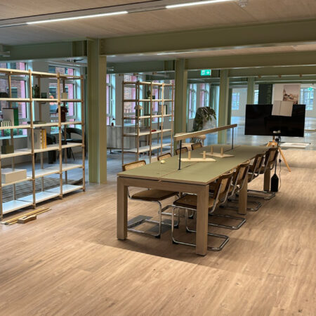 Meeting area with wood-effect flooring, long table and chairs, open shelving and green structural columns.